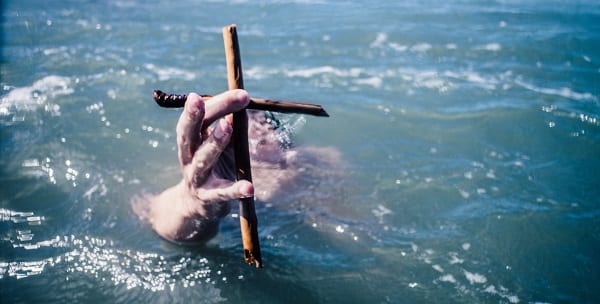 a hand holds a wooden cross of sticks up above the waves